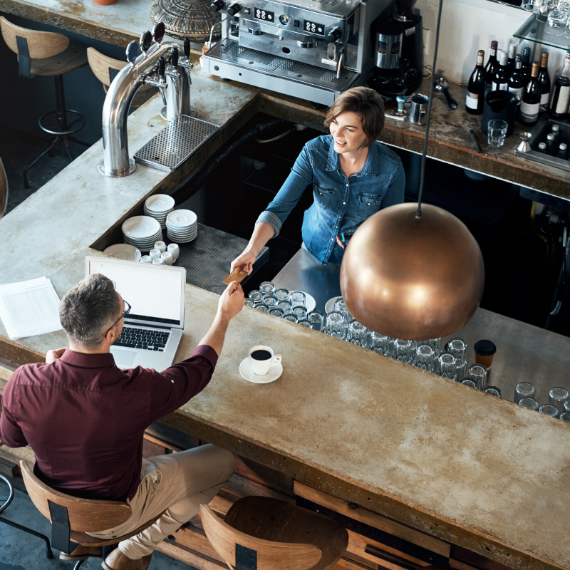 Bird eyes view of a man sitting at a café bar with his laptop and paying the woman behind the counter for his coffee. 